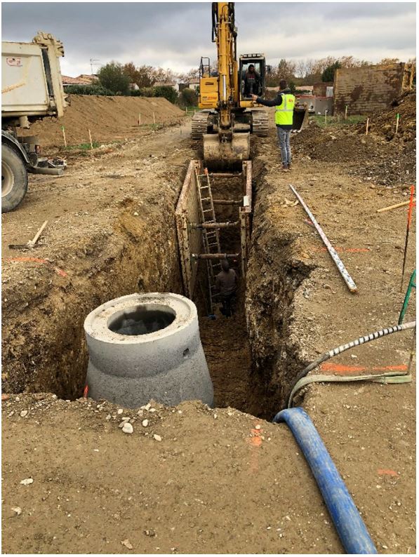 Travaux de terrassement et pose de réseaux enterrés avec engin de chantier et regard en béton dans une tranchée.
